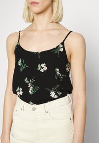 Camisola nera con piccoli motivi floreali in rosa e verde. Spalline sottili regolabili. Abbinata a pantaloni corti bianchi in velluto a coste a vita alta.