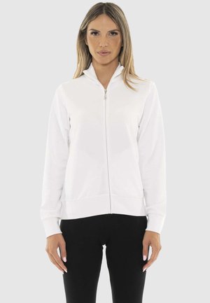 Femme portant une veste blanche zippée à manches longues et un pantalon noir, debout devant un fond clair uni.