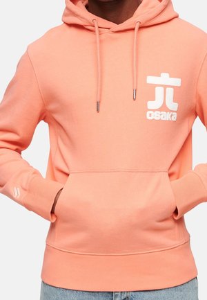 Osoba ubrana w brzoskwiniową bluzę z kapturem z białym logo „osaka” na lewej piersi, ręce w przedniej kieszeni, widoczne niebieskie dżinsy.