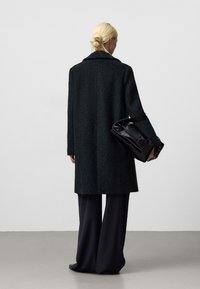 Cappotto verde scuro con un motivo testurizzato, un collo ampio e una vestibilità morbida. Abbinato a pantaloni neri a gamba larga e una borsa nera.