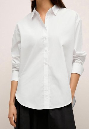 Kvinde iført en hvid skjorte med lange ærmer og knapper, med krave, ærmerne rullet op, og en sort nederdel synlig forneden.