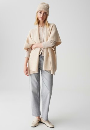 Cardigan in maglia beige con maniche ampie, indossato su un leggero maglione e jeans grigi. Completato da un berretto in maglia coordinato e loafers crema.