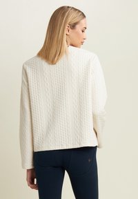Giacca lavorata a maglia color crema con motivi a treccia testurizzati, design cropped e maniche lunghe, abbinata a leggings blu navy.