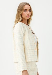 Blonde vrouw in een crèmekleurig, textuur tweedelig pak met gouden knopen, staat zijwaarts en kijkt naar de camera tegen een effen achtergrond.