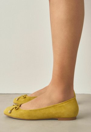 Seitenansicht eines Paares Füße, die senfgelbe Ballerinas mit kleinen dekorativen Schleifen auf einem neutralen Boden tragen.
