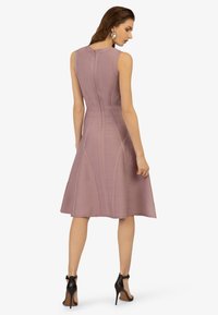 Ärmelloses lilafarbenes Kleid mit enganliegendem Oberteil und ausgestellt Rock. Verfügt über strukturierte, vertikale Linien und einen Reißverschluss hinten. Getragen mit schwarzen High Heels.