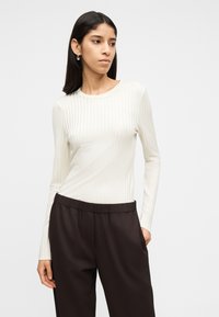 Γυναίκα με μακριά σκούρα μαλλιά που φοράει ασημένια σκουλαρίκια κρίκους, λευκό ριπ μακρυμάνικο τοπ και μαύρο παντελόνι ψηλόμεσο με τα χέρια στις τσέπες.