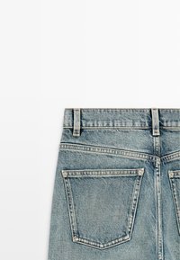 Rückansicht von hellblauen Jeans aus Denim, die den Bund, Gürtelschlaufen und zwei Gesäßtaschen mit Nieten vor weißem Hintergrund zeigen.