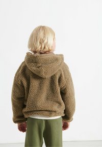 Bambino con i capelli biondi corti di spalle, indossa una giacca cappuccio marrone morbida e pantaloni verdi, sullo sfondo bianco.