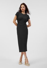 Černé šaty ke kolenům s krátkými rukávy a slim fit, s bočními řasenými detaily. Modelka má na sobě zlaté platformové podpatky. Lehká textura.