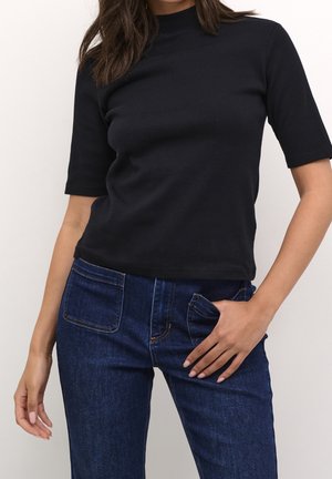 Femme portant un haut noir à manches courtes et col montant et un jean denim bleu foncé avec des poches frontales plaquées, main reposant sur la poche du jean.