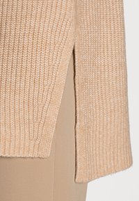 Pull en tricot beige doux avec une texture côtelée, présentant des fentes sur les côtés pour plus de confort. Décoré d'une finition soignée le long des bords.