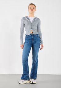 Gray cropped hoodie with ribbed texture and hood, paired with blue flared jeans. Model wears white top and multi-colored sneakers.