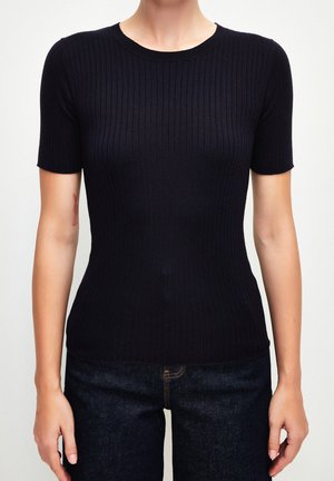 Vrouw draagt een nauwsluitend zwart geribd T-shirt met korte mouwen en donkere jeans, staand met armen ontspannen langs haar zij tegen een effen achtergrond.