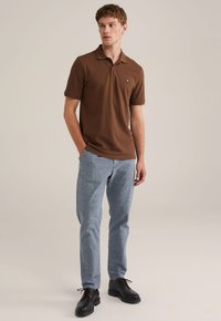 Camisa polo marrón de algodón, con cuello y mangas cortas. Combinada con pantalones de tejido azul claro y zapatos negros de cordones.