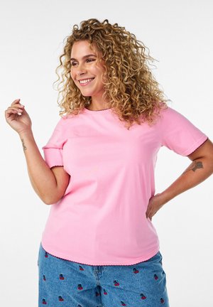 Smilende kvinde med krøllet hår iført lyserød kortærmet skjorte og blå jeans med kirsebærmønster, poserer med hånden på hoften mod en hvid baggrund.
