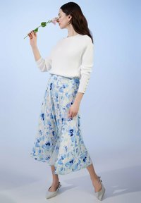 Weißer Pullover mit rundem Ausschnitt und Knopfmanschetten, kombiniert mit einem fließenden, blauen Midikleid mit Blumenmuster und eleganten, hellen Pumps.