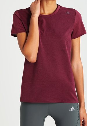 Frau trägt ein eng anliegendes, kurzärmliges maroonfarbenes Adidas-T-Shirt und graue Adidas-Leggings, steht mit einer Hand an ihrem Hals.