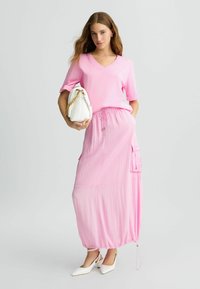 T-shirt rosa a V abbinata a una lunga gonna rosa arricciata con tasche laterali. Borsa bianca con dettagli in catena dorata e scarpe bianche a punta.
