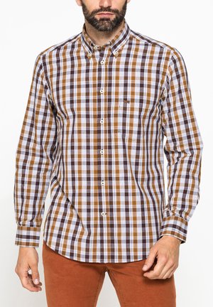 Uomo che indossa una camicia a quadri marrone, bianca e nera con maniche lunghe e pantaloni color ruggine su sfondo bianco.