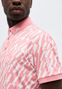 Nærbilde av en person som har på seg en rosa og hvit mønstret polotrøye med rosa krage og "J.LINDEBERG" brodert på ermet.