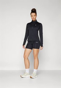 En ung kvinne står i en svart langermet treningsgenser med glidelås, svarte tettsittende shorts, hvite sokker og lyse joggesko.