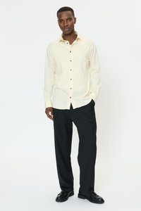 Lichtgeel overhemd met bruine knopen, gedragen met zwarte broek en zwarte schoenen. Het overhemd heeft een kraag en lange mouwen.