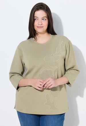 Frau mit langen braunen Haaren, die ein olivgrünes 3/4-Ärmel-Oberteil mit Blumenstickerei und blaue Jeans trägt, steht vor einem weißen Hintergrund.