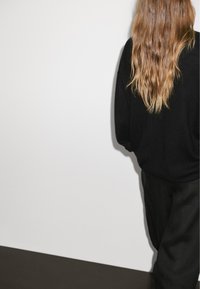 Black sweater with long sleeves, paired with black trousers. Wavy, medium-length hair is visible, against a plain white background.