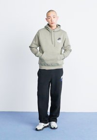 Olivno zeleni kapuzenjak s sprednjimi žepi in črne trenirke z logotipom na levi nogi; kombinirano z belimi in sivimi supergami.