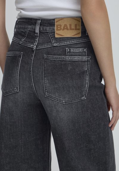 Juodos džinsinės kelnės su aukšta juosmens linija, turinčios išblukusį tekstūrą, dvi užpakalines kišenes ir rudą prekės ženklo etiketę, siuvinėtą gale.