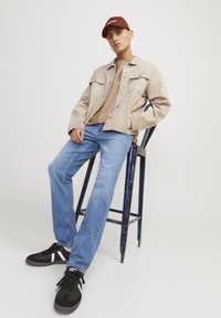 Chaqueta de mezclilla beige sobre una camisa color beige, combinada con jeans azul claro y zapatillas negras con rayas blancas. Modelo sentado en un taburete de madera.