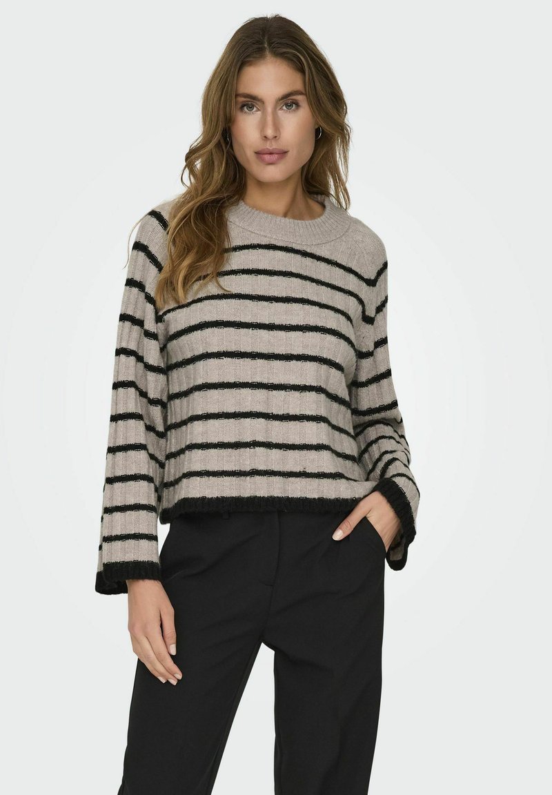 Sweter w paski w delikatnym beżu i czerni, z okrągłym dekoltem i krojem crop, z prążkowanymi mankietami i dołem. Noszony z czarnymi spodniami.