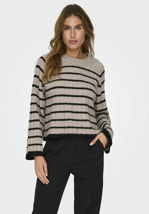 Donna con capelli lunghi e ondulati che indossa un maglione beige con righe orizzontali nere e pantaloni neri, in piedi con una mano in tasca.