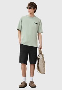 Camiseta de algodón verde claro con texto negro, pantalones cortos de carga negros, bolsa cruzada beige, zuecos marrones, calcetines blancos y gafas de sol.