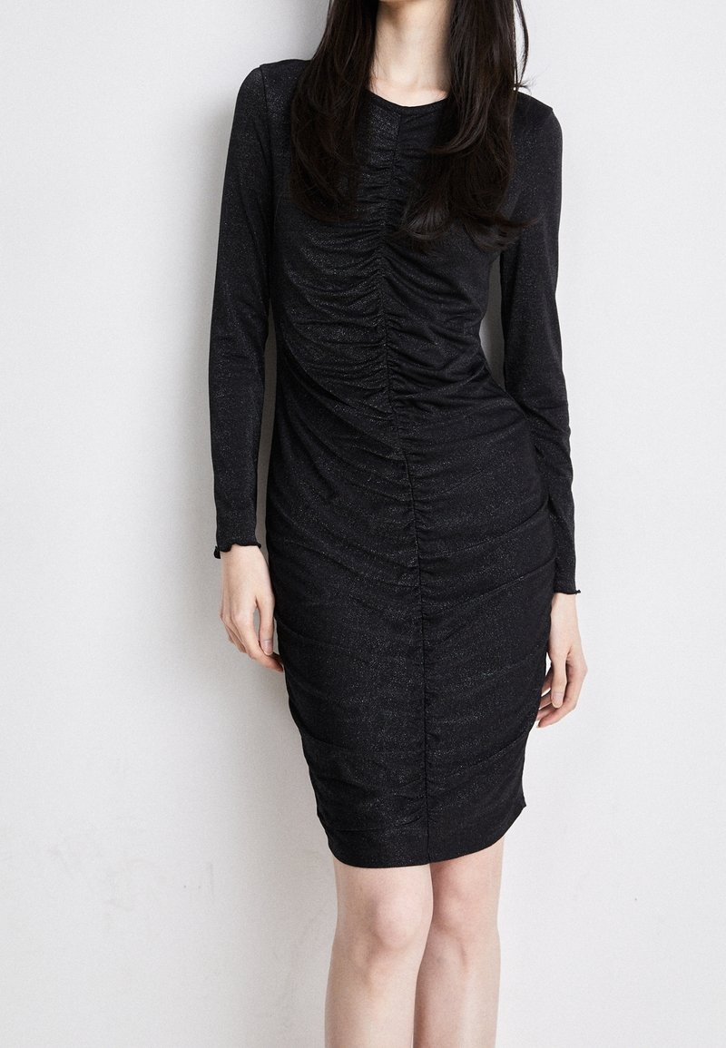 Femme portant une robe noire moulante à manches longues avec des fronces au centre, debout contre un mur uni de couleur claire.