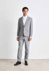 Traje gris con una chaqueta entallada y pantalones rectos, acompañado de una camisa blanca. El material parece suave con un sutil brillo. Zapatos negros.