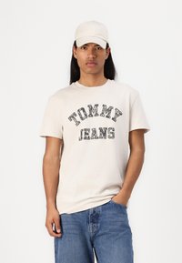 Kremo spalvos medvilnės marškinėliai su juodu užrašu "TOMMY JEANS", derinami su atitinkančiu kremo spalvos kepurėle. Modelis stovi su rankomis kišenėse, vilkėdamas mėlynus džinsus.