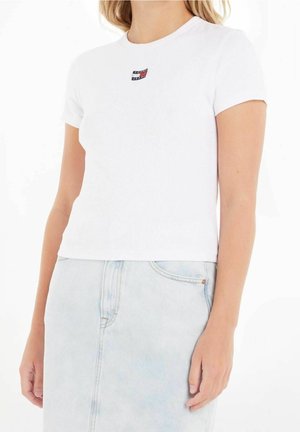 Kvinde iført en hvid crop top med korte ærmer med et lille Tommy Jeans-logo og lyseblå høj talje denimjeans.