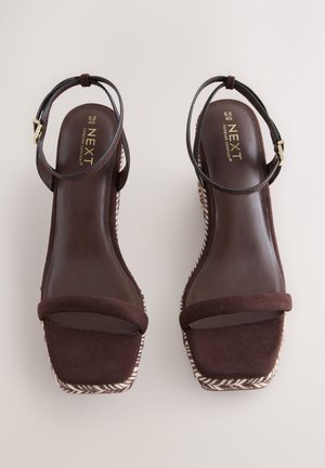 Sandalias cuña marrones con parte superior de ante, correa en el tobillo con hebilla metálica y una plataforma tejida con textura. Plantilla lisa con detalles mínimos.