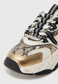 Parte delantera de zapatilla gruesa con detalles en blanco, negro, dorado y estampado de serpiente, con cordones gruesos a rayas negras y blancas y etiqueta de metal con la marca en el cordón.