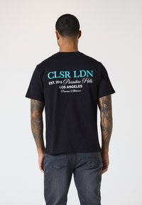 Svart bomull t-skjorte med turkise og hvite bokstaver på baksiden som viser "CLSR LDN," "Paradise Hills," og "Los Angeles."