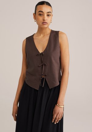 Brown linen sleeveless top with a deep V-neck, front ties, and a slightly curved hem, paired with a black skirt. Smooth, textured fabric.