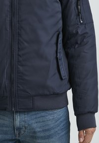 Chaqueta bomber azul marino con cierre frontal, puños y cinturilla acanalados. Cuenta con un bolsillo en el brazo izquierdo y una textura lisa y resistente al agua.