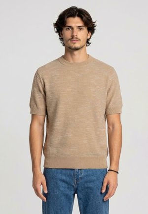 Uomo con capelli scuri e barba che indossa un maglione beige a maniche corte lavorato a maglia e jeans blu su sfondo chiaro uniforme.