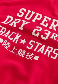 Červené bavlněné tričko s odvážným, popraskaným bílým nápisem "SUPER DRY 23" a "TRACK STARS" spolu s dalšími znaky v menším písmu.