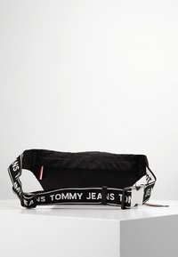 Svart tygmidjeväska med ett brett justerbart band som har upprepande texten "TOMMY JEANS", silverfärgad spänne och subtila logodetaljer.