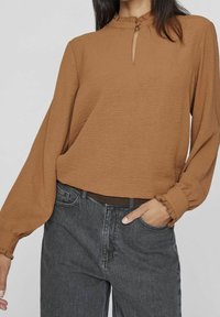 Brun krinklad långärmad topp med hög hals, nyckelhålsdetalj och volangärmar, kombinerad med mörkgrå högmidjade jeans.