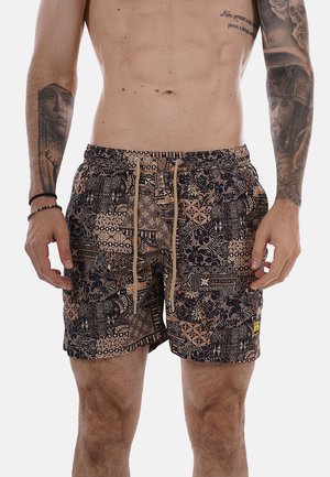 Torace maschile con tatuaggi su entrambe le braccia che indossa pantaloncini marroni e neri con coulisse, su uno sfondo semplice.