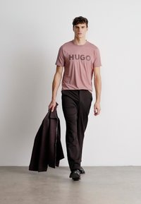 Modelka oblečená v mäkkej ružovej tričku s nápisom "HUGO" v tmavých písmenách, skombinovaná s čiernymi nohavicami, drží tmavú bundu a stojí na betónovej podlahe.